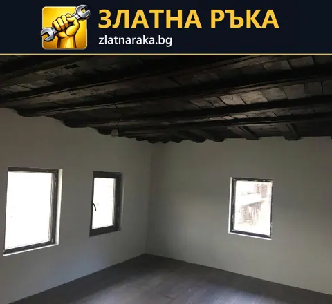 Празна стая с бели стени, три прозореца, тъмни таванни греди и сива подова настилка