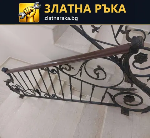 Кафяв дървен ръкохват върху тъмен метален парапет с извивки над светли вътрешни стъпала