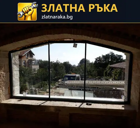 Голям прозорец с три вертикални полета и тъмна рамка под дъговидна тухлена арка в грубо помещение
