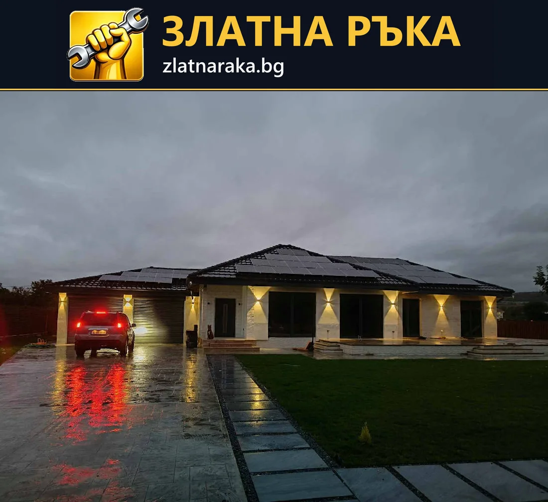 Осветена къща с тъмен покрив, гараж и мокра дворна настилка, отразяваща светлината от фасадата