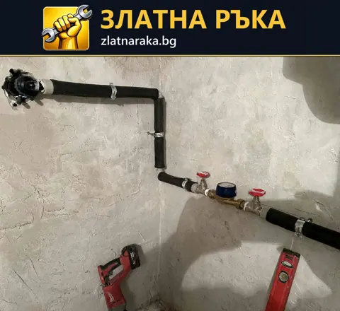 Открит водомерен възел с два червени крана и тъмни тръби върху необработена стена