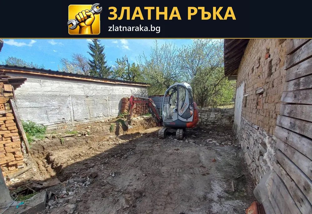 Малък багер върху разкопан терен между тухлена постройка и оградна стена.