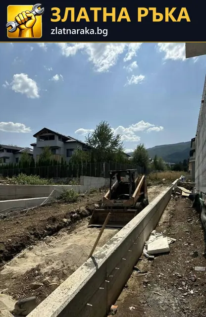 Малка строителна машина в изкоп до дълга бетонна стена и незавършена дворна площ