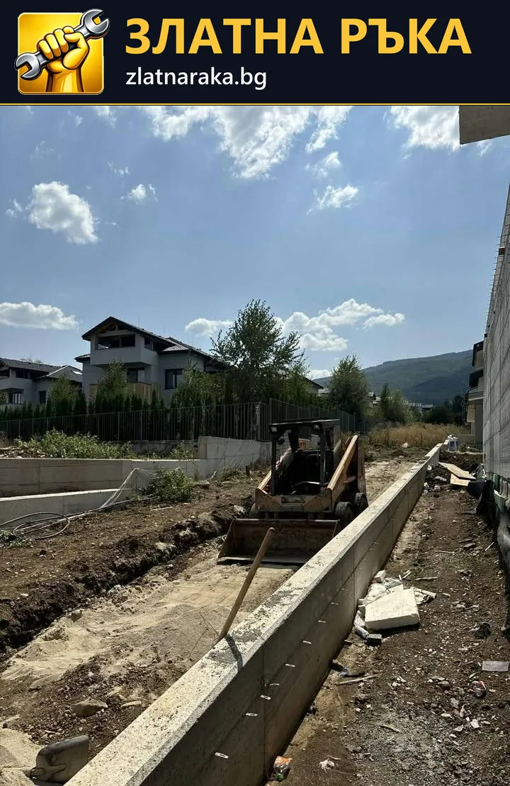 Малка строителна машина в изкоп до дълга бетонна стена и незавършена дворна площ