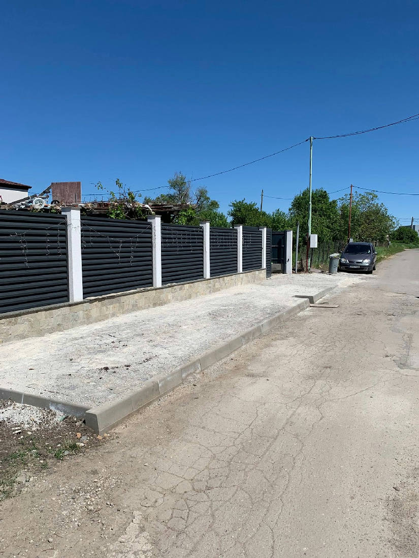 Тротоарна площ с нов бордюр и светла настилка, засипана с пясък, покрай ограда с тъмни метални ламели и бели колони.